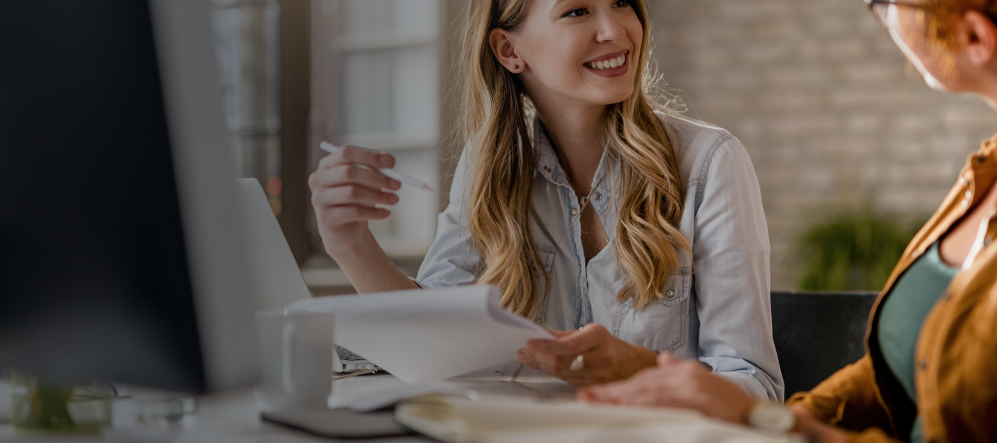 smiling-creative-businesswoman-going-through-paperwork-talking-her-female-colleague-office 1